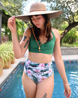 Woman in green bikini top and floral bottoms, wearing a wide-brimmed honey palm straw hat with leather chinstrap by a pool. - model