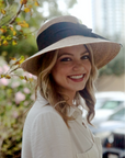 Smiling model wearing a Joliet straw hat with an asymmetrical brim and black ribbon bow.