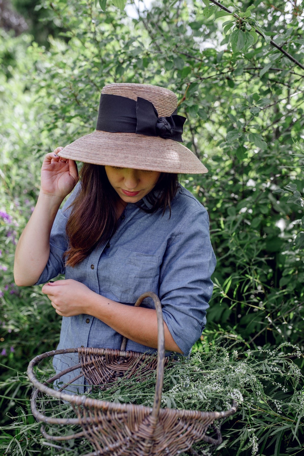Model in a wide-brimmed natural palm straw hat with a black band and bow, harvesting in a garden.