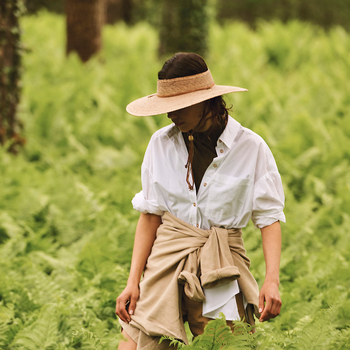 Hand-woven natural palm straw open crown hat with leather chinstrap, worn by model in a fern forest.