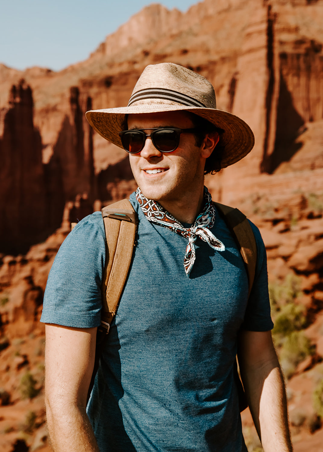 Man hiking in a Clark Straw Hat with brown band, sunglasses, and backpack, against red rock canyons - model